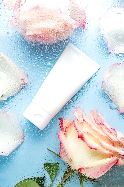 Cosmetic tube for face cream cleanser or body lotion on a blue background with water droplets and rose flowers through the glass The concept of a moisturizing cosmetic product