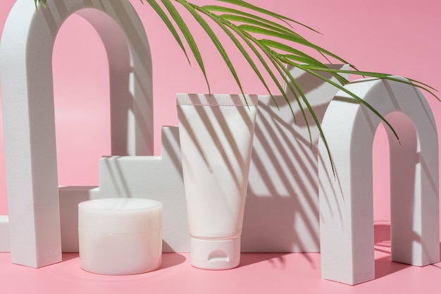 Cosmetic white containers with palm leaf on pink background. Skin care products. Hand, face, body moisturizing cream jar and plastic tube.