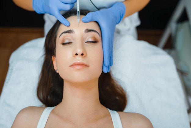 Cosmetician in gloves gives face botox injections to female patient on treatment table.