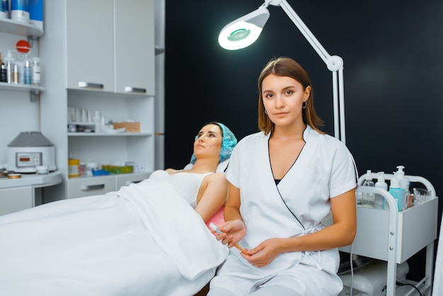 Cosmetician with brush sitting near female patient on treatment table. Rejuvenation procedure in beautician salon. Doctor and woman, cosmetic surgery against wrinkles and aging