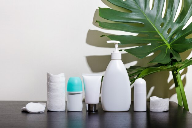 Cosmetics in white tubes and bath products on a black table with tropical leaves.