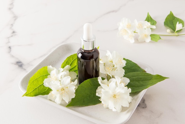 Cosmetische jasmijnolie in een glazen fles op een keramisch dienblad met verse bloemen zorg voor de huid van het gezicht en lichaam marmeren witte achtergrond