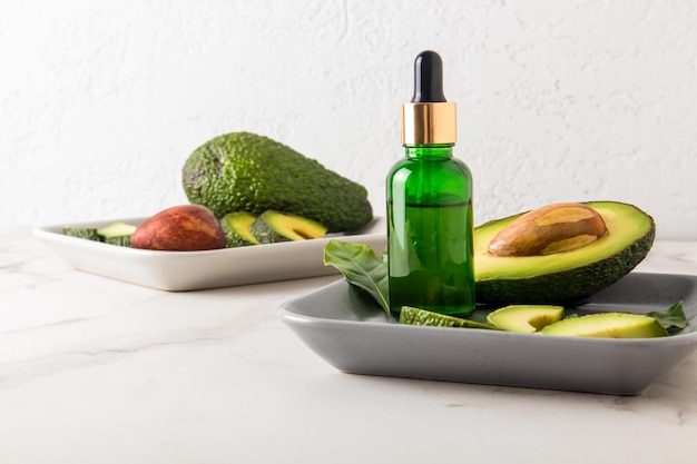 cosmetische olie met avocado-extract in een fles met een druppelaar op een keramische rechthoekige plaat met avocadofruit tegen een witte muur.