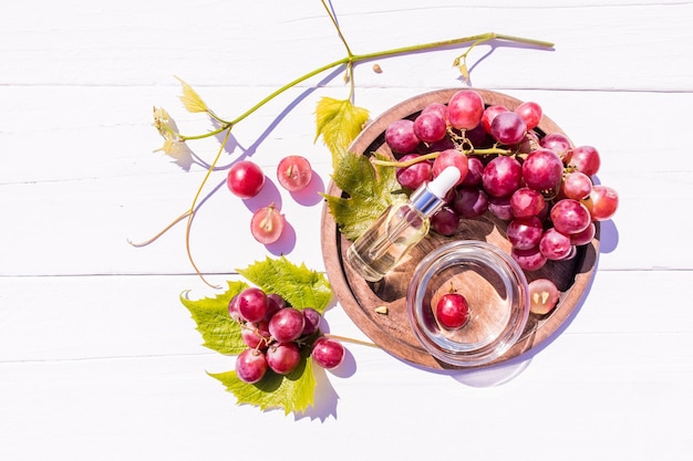 Cosmetische olie wei op basis van druivenpitolie in een glazen flesje met een pipet op een houten dienblad met een tros druiven bovenaanzicht witte houten achtergrond