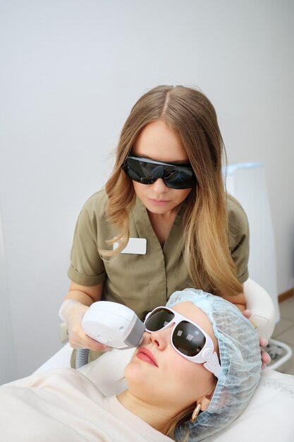 Cosmetological clinic worker performs radical hair removal on girl patient face using improved special device