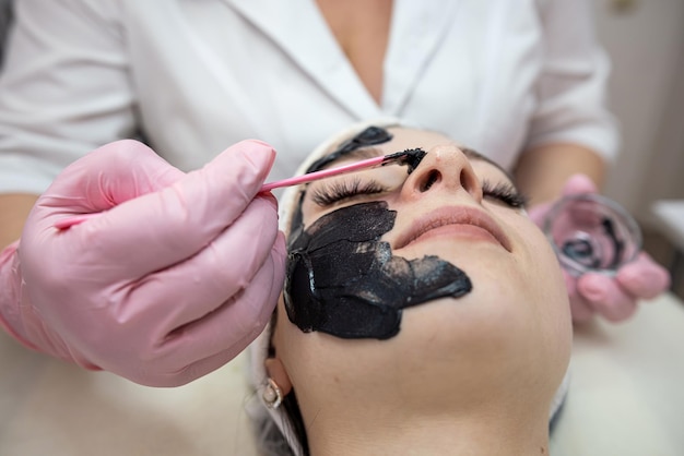 Cosmetologist applying black cream mask on face her client