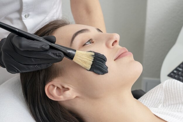 Cosmetologist applying black mask on the face of a beautiful woman for carbon peel