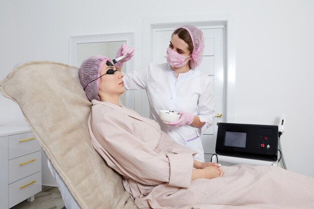 Cosmetologist applying black mask on the face of a beautiful woman for carbon peeling