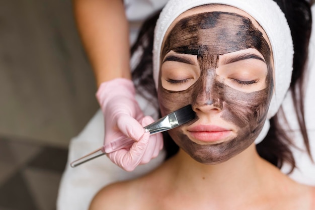 Cosmetologist applying black mask on pretty woman face wearing\
black gloves gorgeous woman in spa having facial procedures