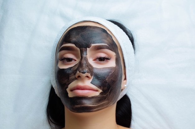 Cosmetologist applying black mask on pretty woman face wearing black gloves gorgeous woman in spa