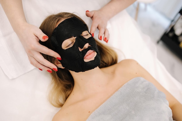 Cosmetologist applying black moistuizing mask on womans face in beauty salon beauty procedure for