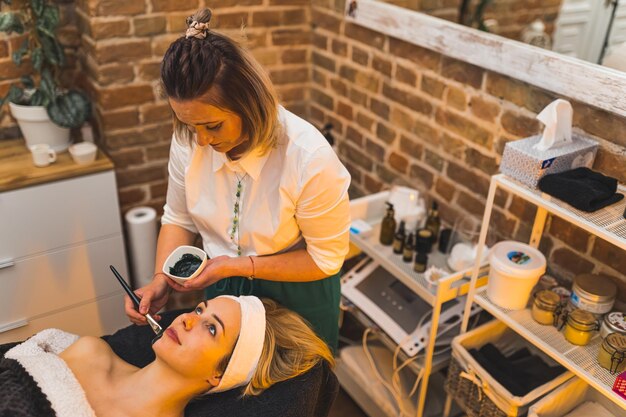 Cosmetologist applying charcoal mask using a brush to her client in a beauty spa