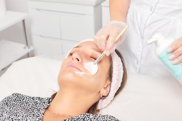 Cosmetologist applying cosmetic cream mask on female face for rejuvenation procedure in beauty salon