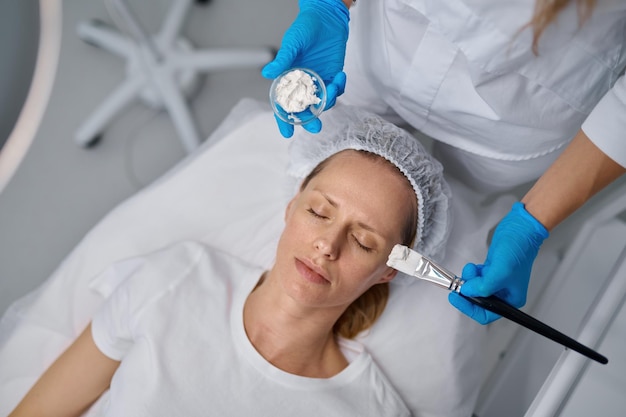 Cosmetologist applying mask on client face using brush. Professional cosmetology and beauty treatment in spa salon