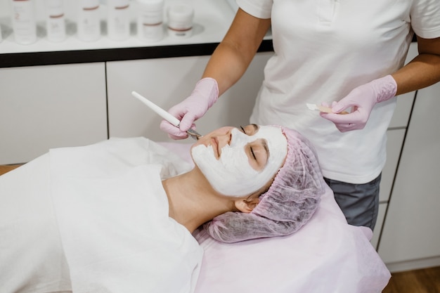 Cosmetologist applying mask on clients face in spa salon young woman getting facial care by