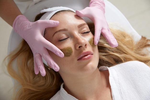 Cosmetologist applying a mask from clay to the face of a beautiful woman spa treatment and face care