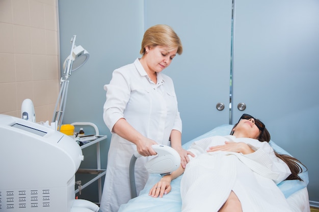 Cosmetologist beautician with laser hair removal equipment, body and skin care concept. Young woman remove hair on armpits at cosmetology clinic.