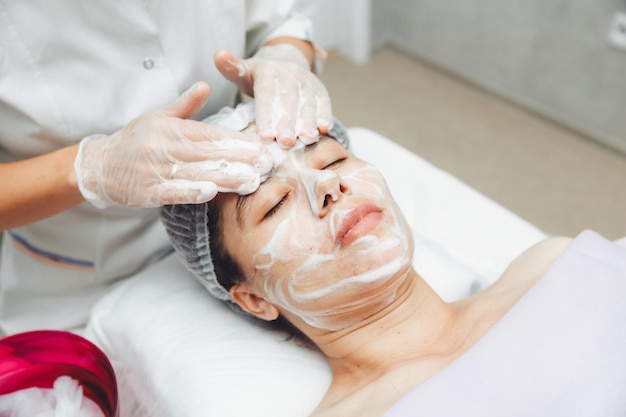 The cosmetologist cleanses the skin with foam A woman's skin care procedure Natural cosmetics A beautiful face Cosmetic treatment of the face Therapeutic peeling cream