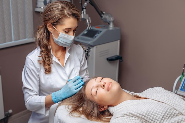 A cosmetologist conducts mesotherapy for the hair of a young woman lying on a white couch in the office