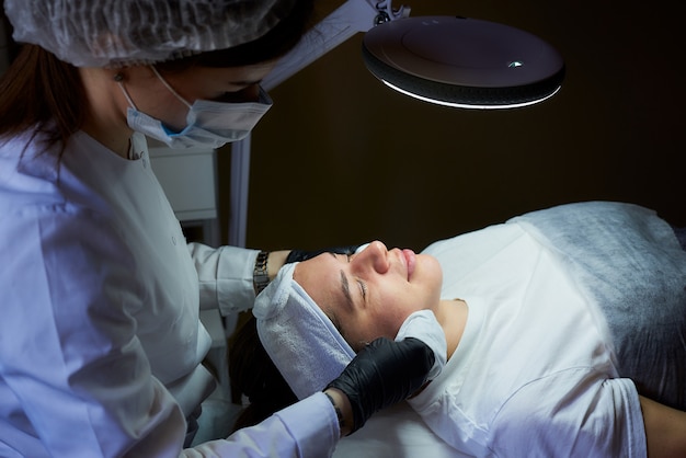 Cosmetologist die een chirurgisch gezichtsmasker en beschikbare medische handschoenen draagt reinigt met servetten het gezicht van een vrouw