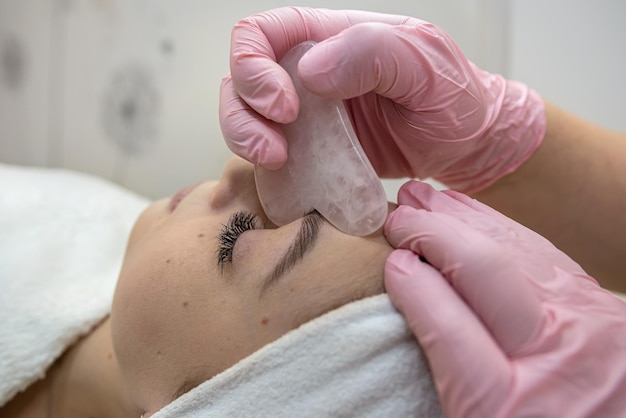 Косметолог выполняет медицинскую процедуру с помощью белого скребка гуаши на лице клиента