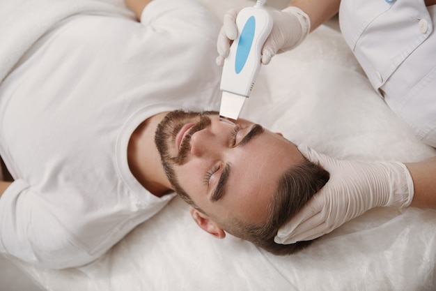 Косметолог очищает поры кожи своим клиентом-мужчиной с помощью ультразвука