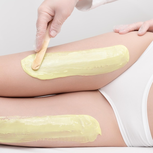 Cosmetologist in gloves applies hot wax on slim woman legs using spatula Depilation in beauty salon