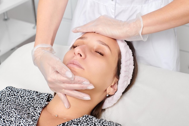 Cosmetologist in gloves massages female face skin for rejuvenation procedure in beauty salon