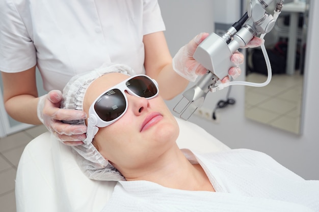 Cosmetologist hand holds neodymium laser at patient face in glasses doing cosmetological procedure selective focus