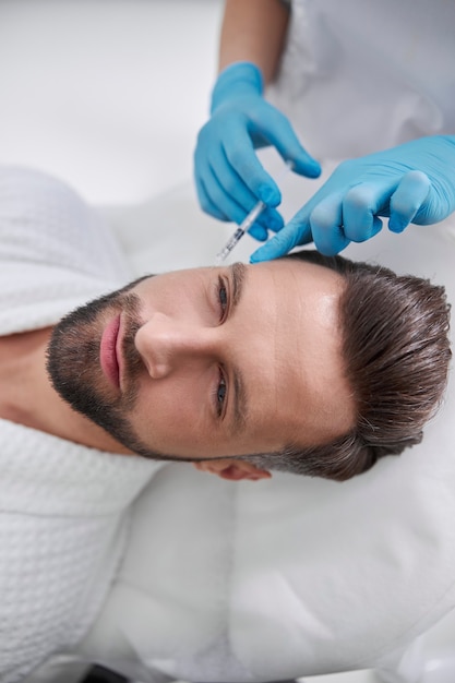 cosmetologist holds finger on mature man client temple after injection of filler for skin lifting