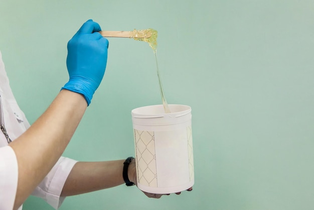 The cosmetologist holds a jar of sugar wax shows texture of paste with a spatula