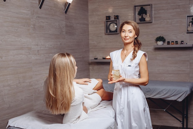 The cosmetologist is holding a glass of cold water with lemon and is going to give it to a woman in a white coat lying on the couch