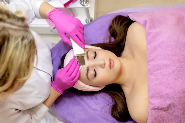 The cosmetologist makes the procedure ultrasonic face peeling of the facial skin of a beautiful, young woman in a beauty salon.