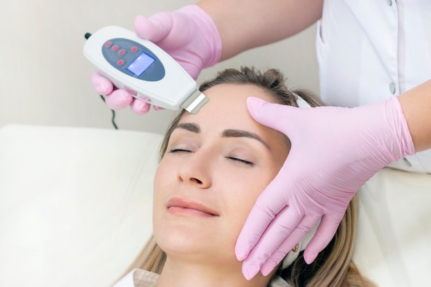 Cosmetologist makes an ultrasonic cleaning of the face of a young woman.