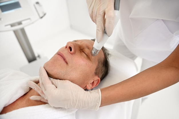 Cosmetologist making procedure of the face skin of a handsome middle aged 50 years old in a beauty salon. Cosmetology and professional skin care concept in wellness spa. Modern hardware cosmetology