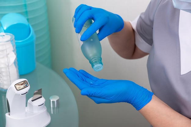 医療用マスクと灰色のコートを着た美容師が、青い使い捨て手袋をはめてスムージングトナーを手に注ぎます。