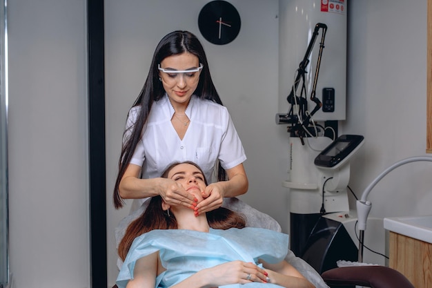 A cosmetologist in protective glasses stands over a client lying on a chair and does an anti-aging facial massage.