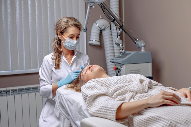 A cosmetologist in a protective mask and gloves performs a cosmetology procedure for a woman in a bathrobe lying on a couch