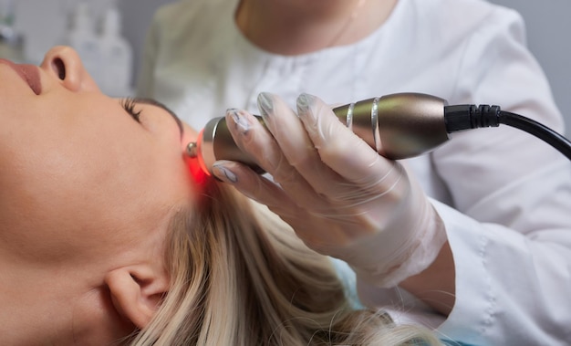 A cosmetologist in rubber gloves does the rflifting procedure on the face In the background the Rf cosmetology device Close up Side view The concept of professional cosmetology and rejuvenation