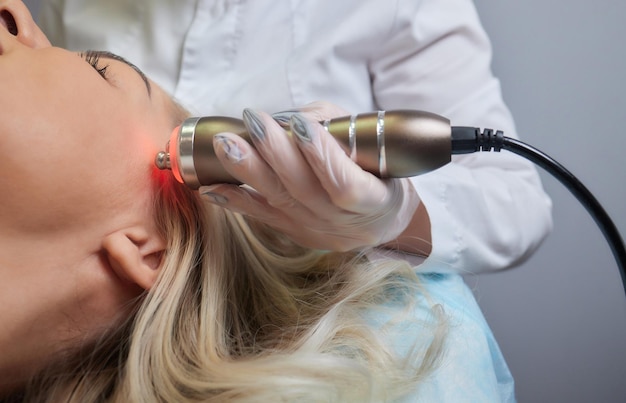 A cosmetologist in rubber gloves does the rflifting procedure on the face In the background the Rf cosmetology device Close up Side view The concept of professional cosmetology and rejuvenation