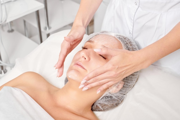 Cosmetologist rubbing moisturizing cream mask into woman face skin for rejuvenation in beauty salon