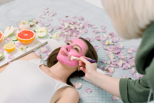 Cosmetologist smeert cosmetische gezichtsmasker op het gezicht van de mooie jonge vrouw in de spa salon. Gezichtsmasker, spa schoonheidsbehandeling, huidverzorging.