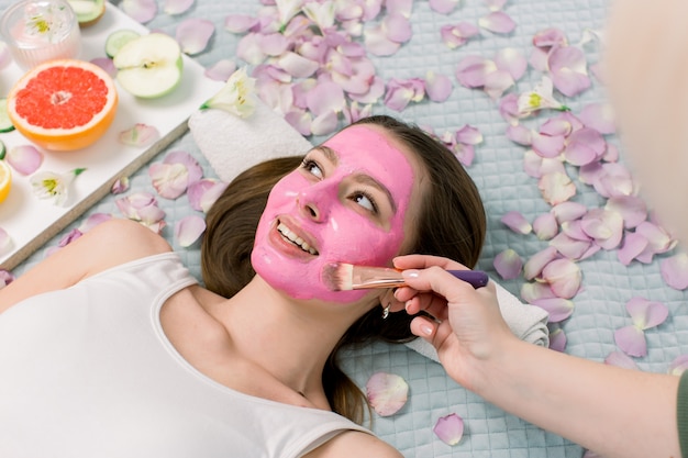 Cosmetologist smeert cosmetische gezichtsmasker op het gezicht van de mooie jonge vrouw in de spa salon. Gezichtsmasker, spa schoonheidsbehandeling, huidverzorging.