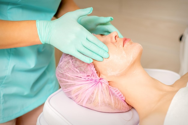 Cosmetologist with gloved hands applies a moisturizing mask with peeling cream on the female face Facial cosmetology treatment Procedures for facial care