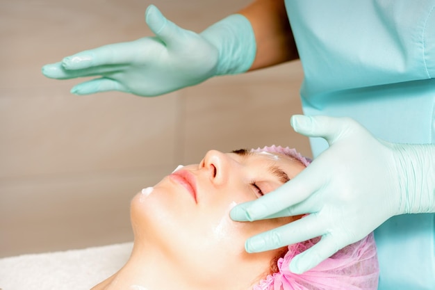 Cosmetologist with gloved hands applies a moisturizing mask with peeling cream on the female face Facial cosmetology treatment Procedures for facial care