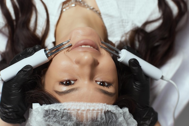 Cosmetology. Beautiful Woman At Spa Clinic Receiving Stimulating Electric Facial Treatment From Therapist.