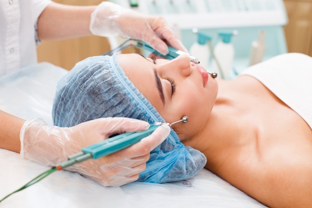 Cosmetology. Beautiful Woman At Spa Clinic Receiving Stimulating Electric Facial Treatment From Therapist.