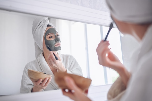 Cosmetology, skin care, face treatment, spa, natural beauty concept. Beautiful smiling woman at home in bathrobe with a towel applying face clay mask against acne to rejuvenate problem skin