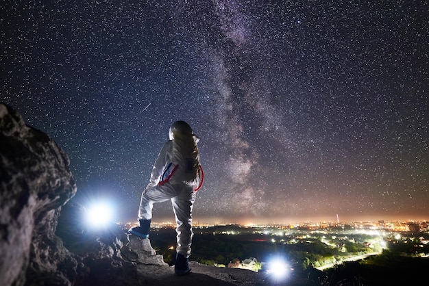 岩が多い丘の上に立って、夜空を見ている宇宙飛行士