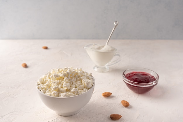 Cottage cheese in a bowl, sour cream with spoon and fruit jam on a light background.